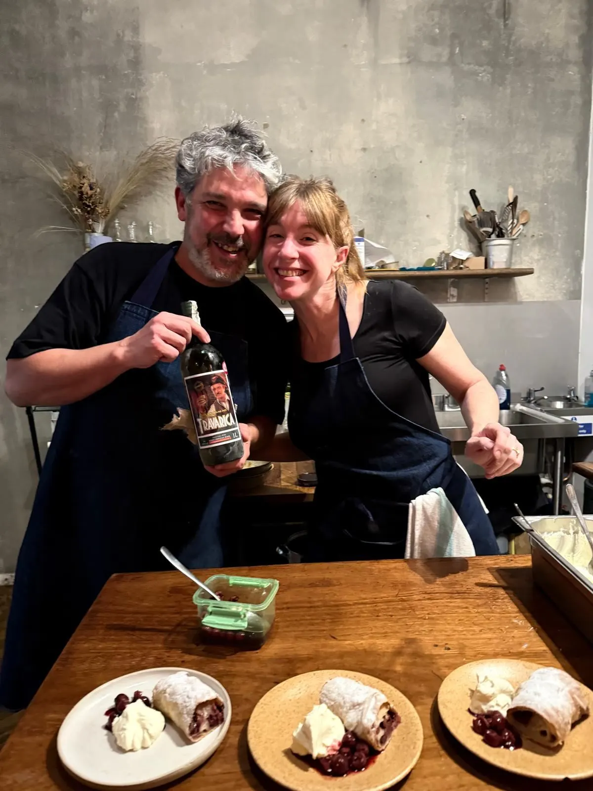 Two cooks in aprons smiling and holding a bottle of travarica over plates of strudel