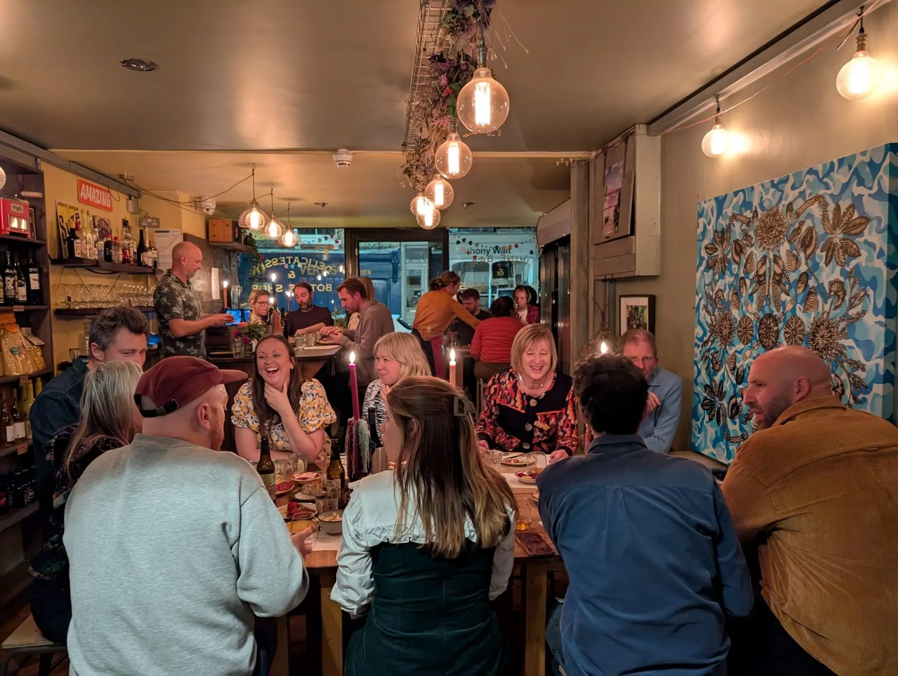 A long candlelit table of guests laughing in a small wine bar